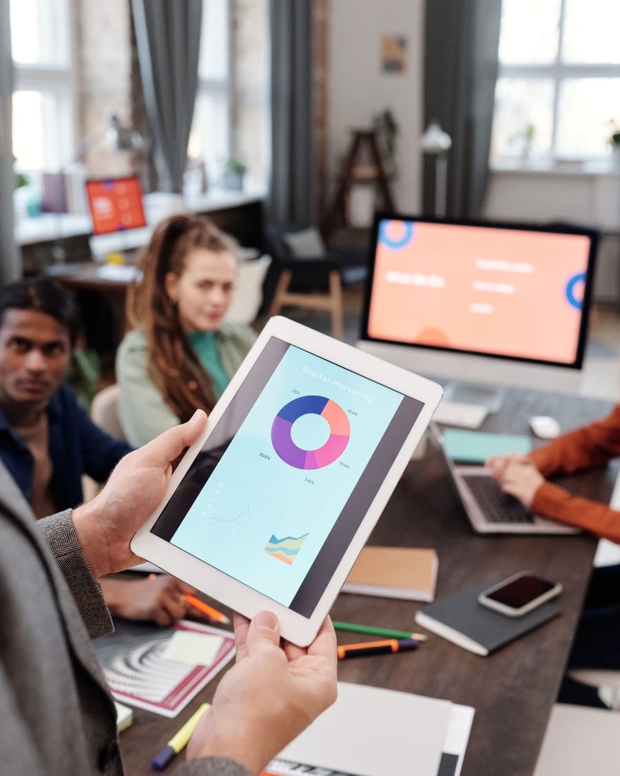 A client holding a tablet showing a digital marketing dashboard, colleagues at the table behind