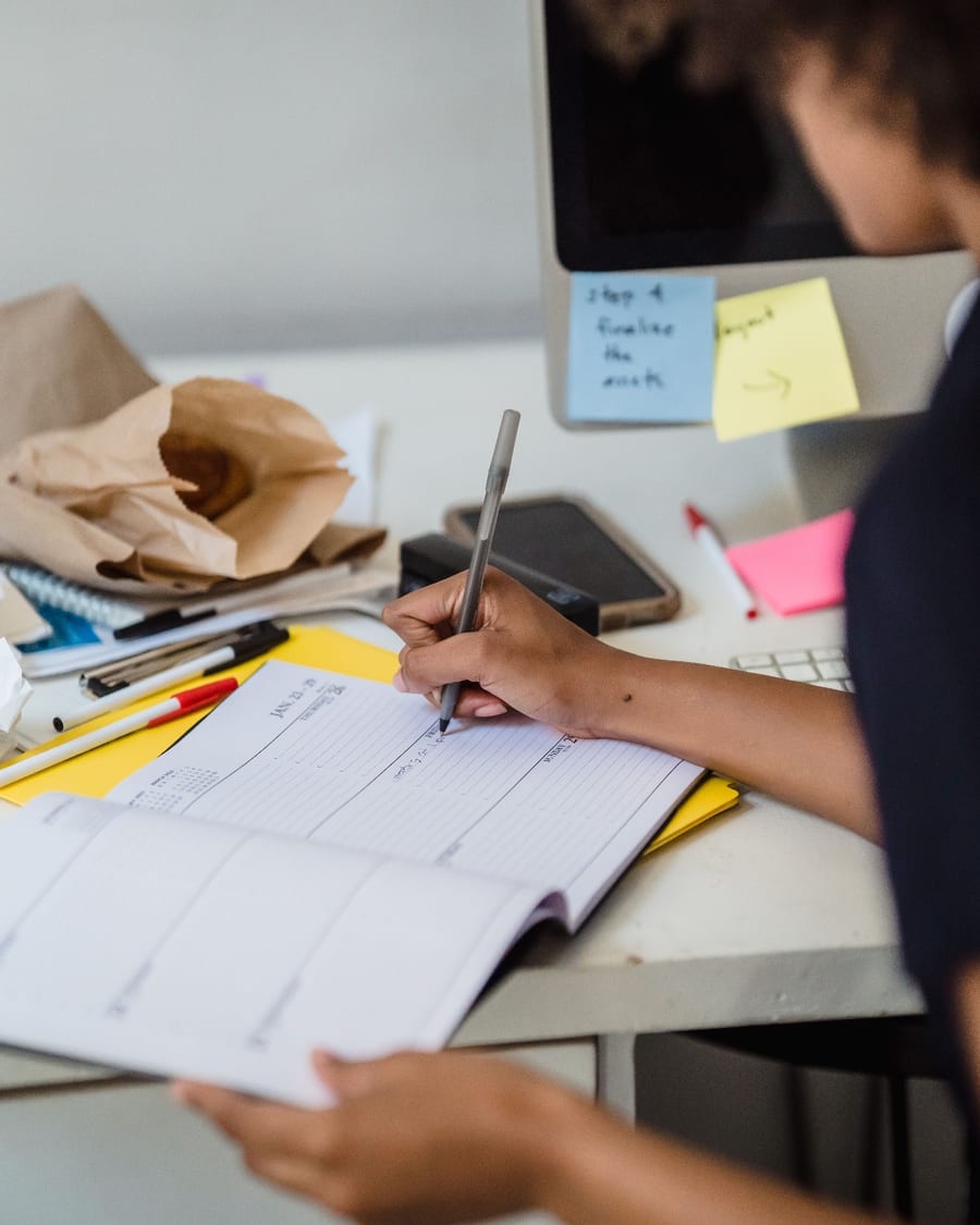 A marketing lead writing the week's plan into a paper planner beside a monitor of sticky notes