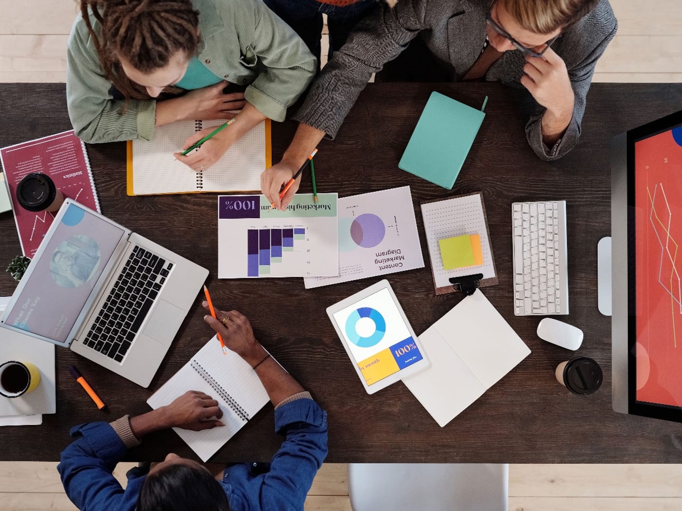 Three people around a shared desk, seen from above, with marketing charts, notebooks and a laptop between them